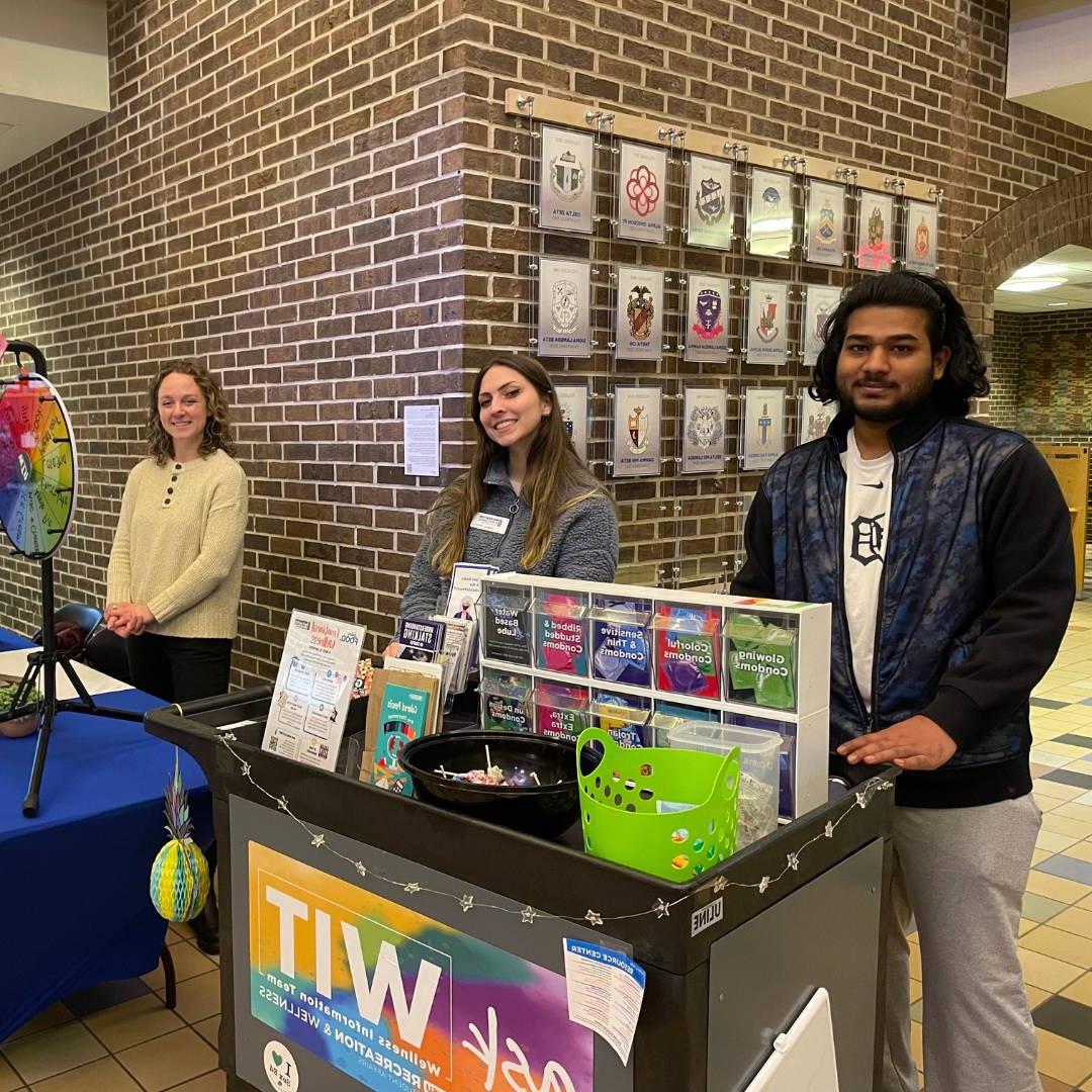 Image of two WIT Peer Educators tabling in Kirkhof behind the WIT cart and the on-campus registered dietitian behind a spinning wheel